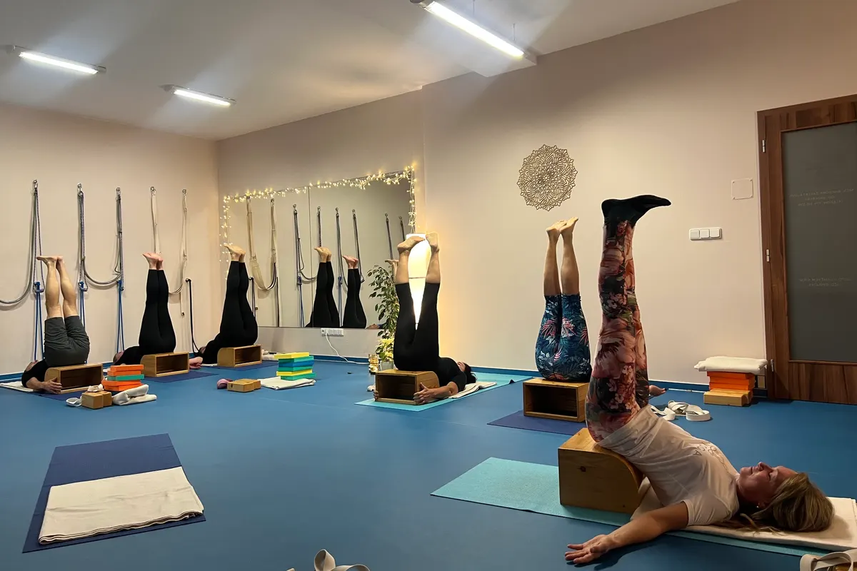 Skupina cvičících ve studiu TaYoga s nohama vzhůru a pánví na dřevěných bednách, vzadu zrcadlo se světýlky